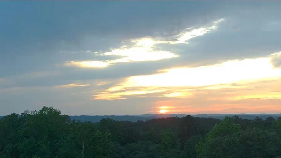 Sunset over central Alabama, the sun dropping behind low forested ridges under streaked orange and gray clouds.
