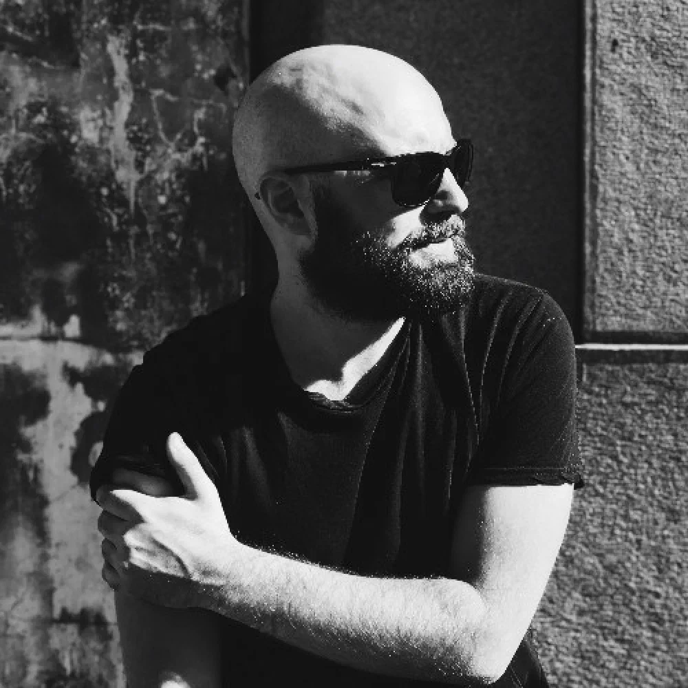 Black-and-white profile photo of Chris Macho, a bearded, bald creator in sunglasses and a black t-shirt against a textured concrete wall.