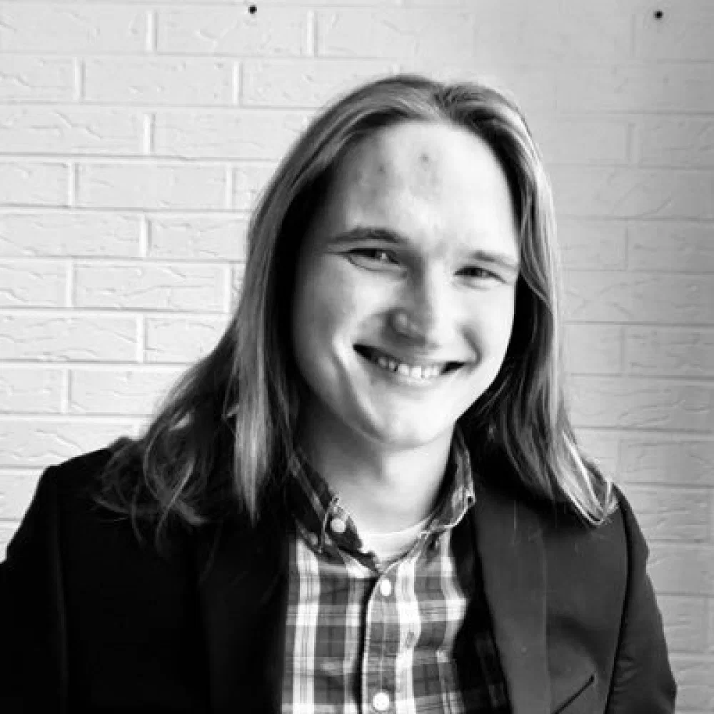 Black-and-white headshot of Josh Lambert, a young man with long blonde hair smiling in a button-up plaid shirt and blazer against a white brick wall.