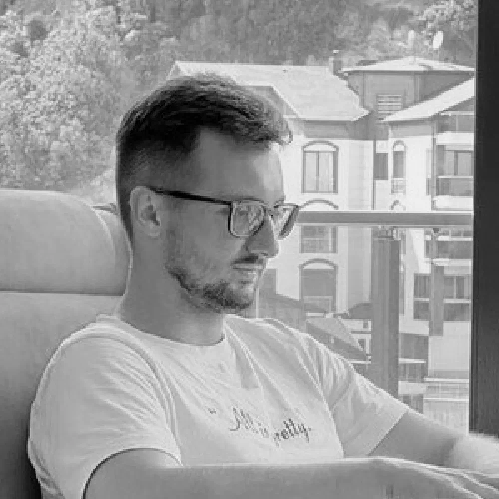 Black-and-white window-light photo of Andrei Zobnin, a bearded young creator in glasses, looking off-frame from a balcony with a hillside village beyond.