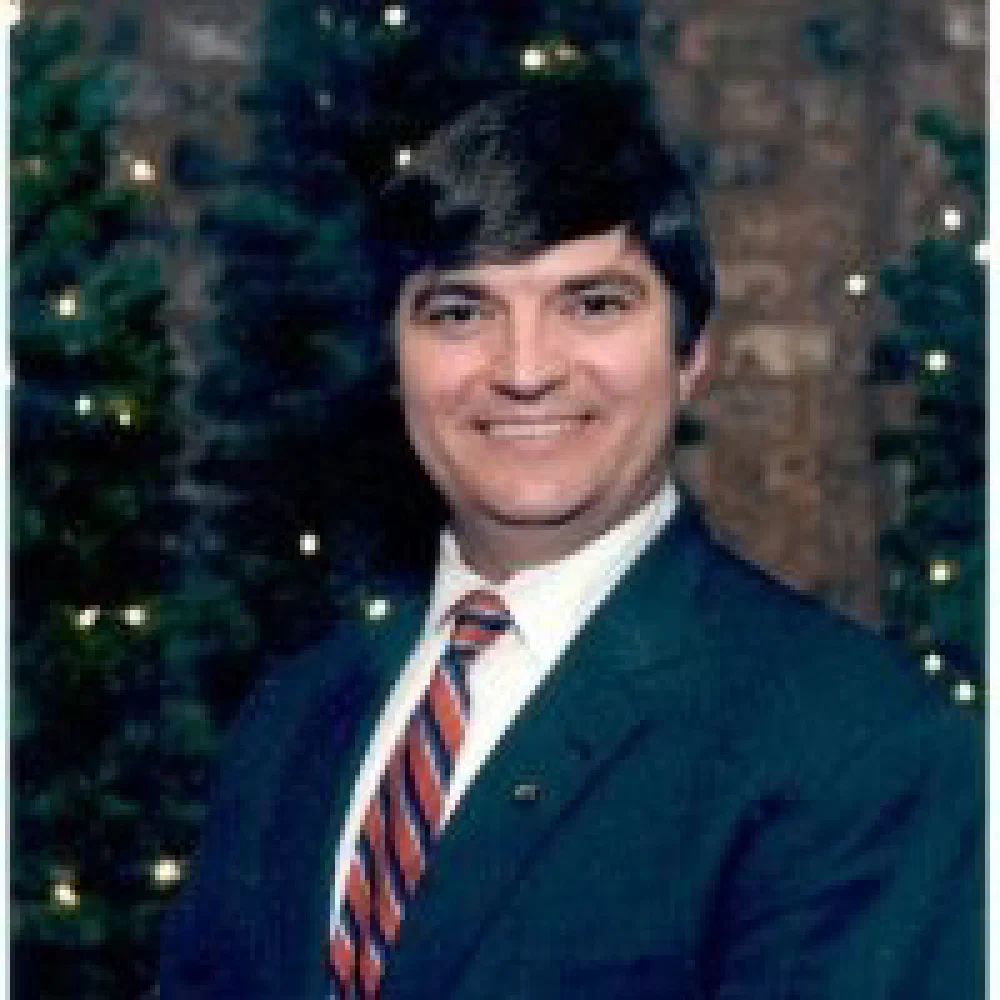 Studio portrait of an older man in a dark green sport coat, striped tie, and red lapel pin posing in front of a Christmas tree.