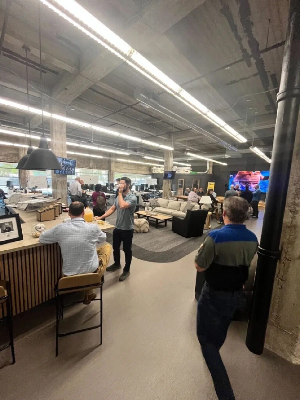 Wide angle of the Birmingham DBA event space, with attendees gathered along a bar and lounge area while a video wall plays surveillance demos in the background.