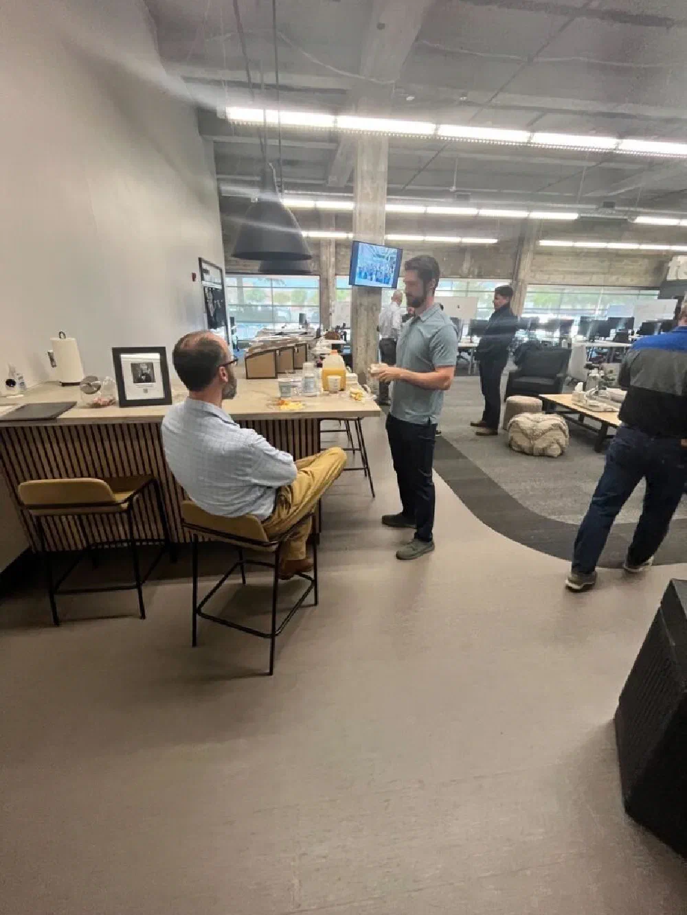 Two men in casual business attire chatting at a wooden bar inside the DBA event space at Innovation Depot, with monitors and breakfast spread in the background.