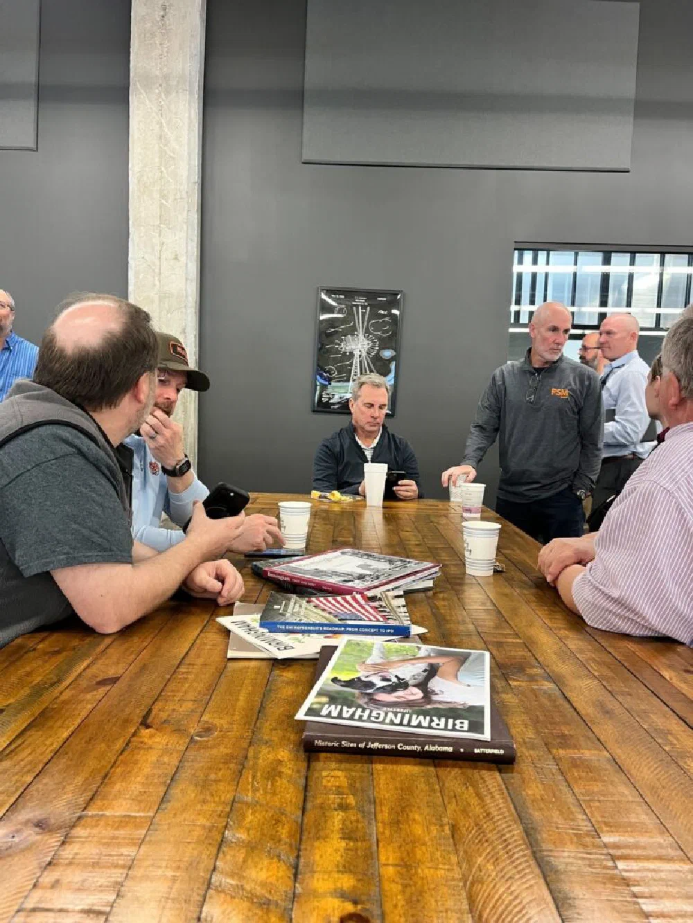 DBA event attendees seated around a wooden table with takeaway coffee cups and Birmingham history coffee-table books, an FSM-branded organizer standing to greet a newcomer.