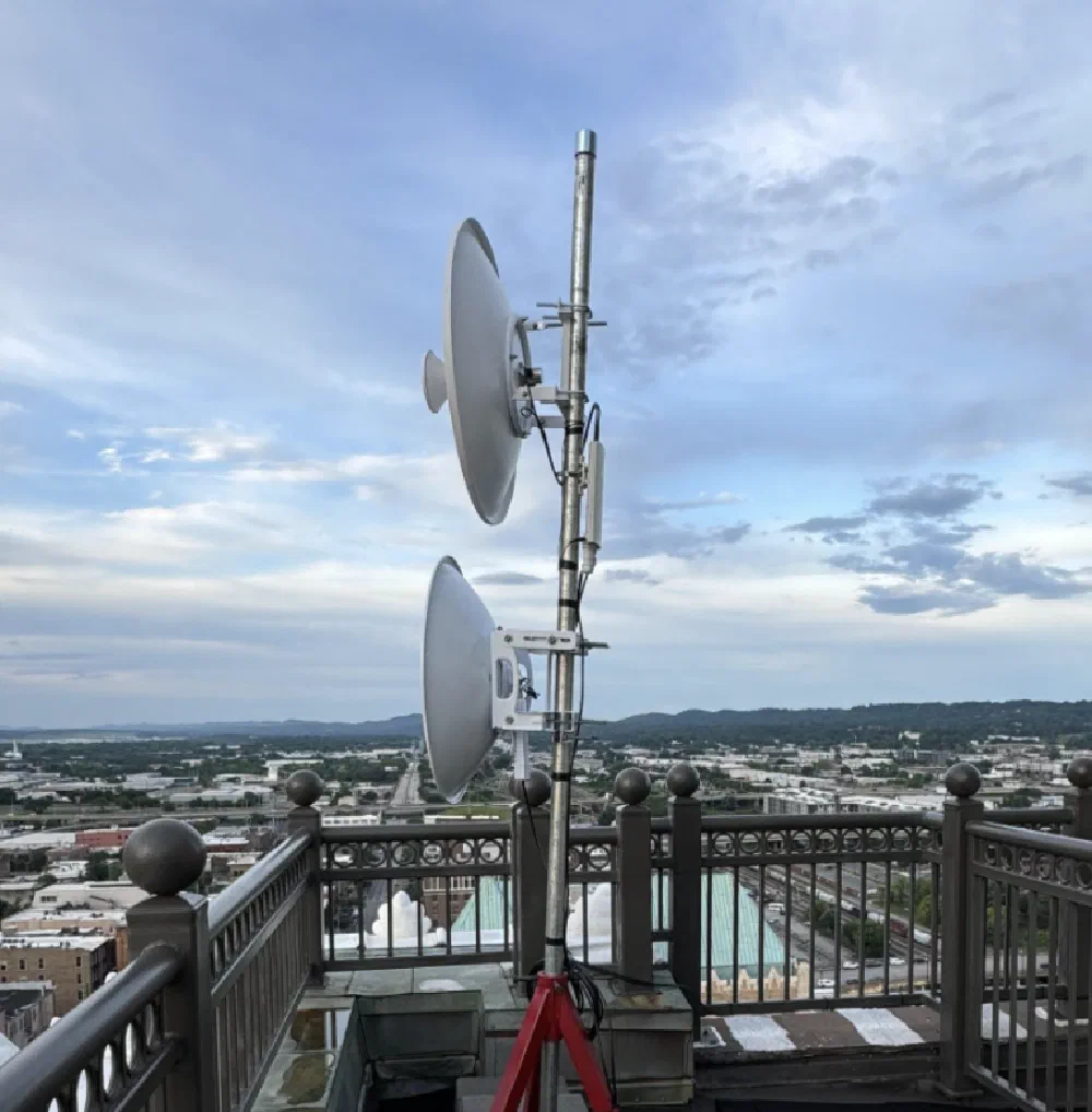 Pair of parabolic backhaul radios mounted on a rooftop balcony rail above a downtown Birmingham skyline view at golden hour.