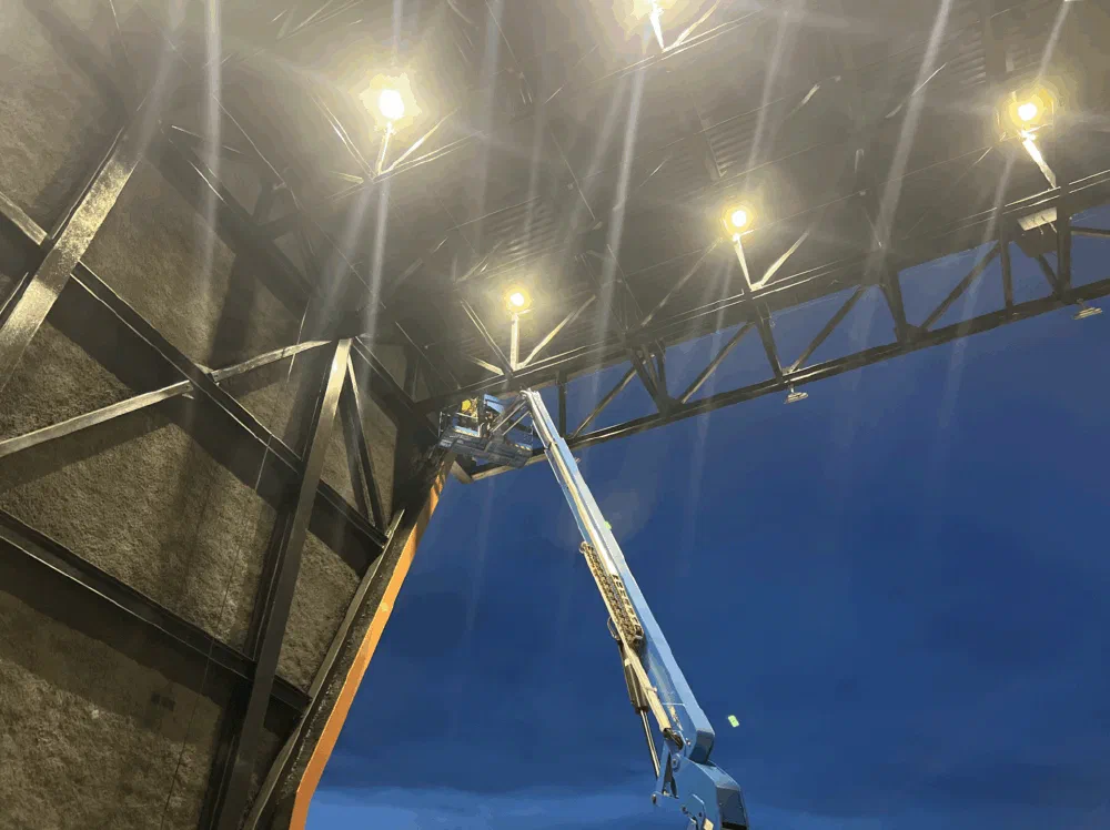 Articulated boom lift extended toward the rafter trusses inside the Birmingham Amphitheater at night, lit by overhead floodlights.