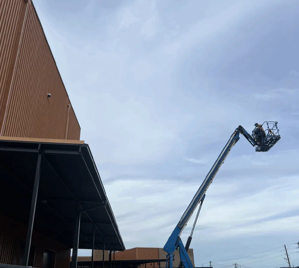 Blue Genie articulated boom lift extended toward the corrugated-metal exterior of the Birmingham Amphitheater on a cloudy afternoon.