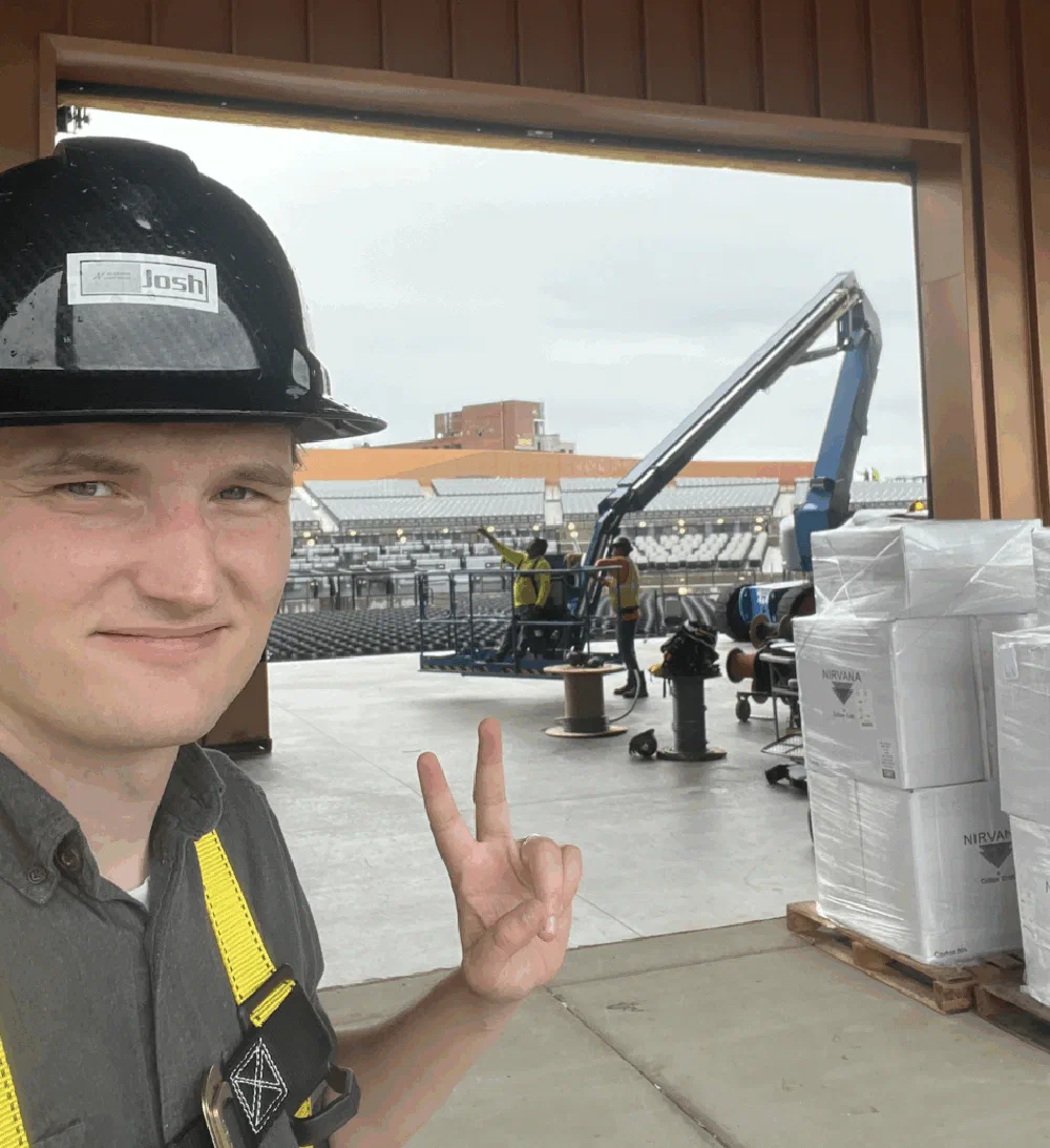 Young Alabama Lightwave technician in a hard hat flashing a peace sign inside an open warehouse loading dock at the amphitheater build site.