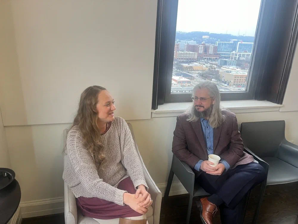Two TiDev attendees, a long-haired woman and a long-haired man in a maroon blazer, seated in armchairs talking in front of a window overlooking downtown Birmingham.