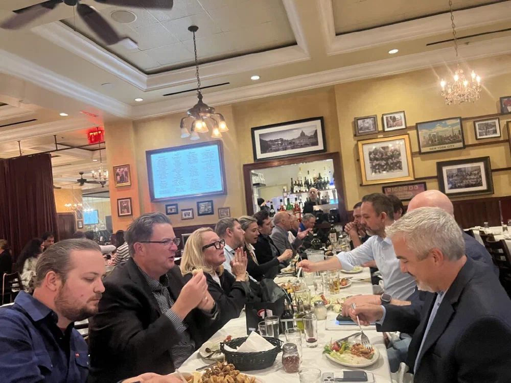 Group dinner at a New York Italian restaurant with hammered-tin ceiling and chandeliers, with attendees in business attire passing dishes around a long white-cloth table.