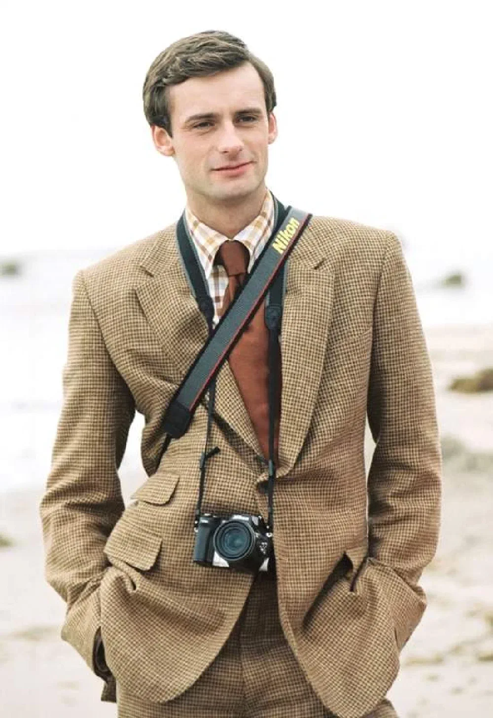 Period-style portrait of a young man in a tweed houndstooth suit on a beach, with a Nikon camera slung around his neck.