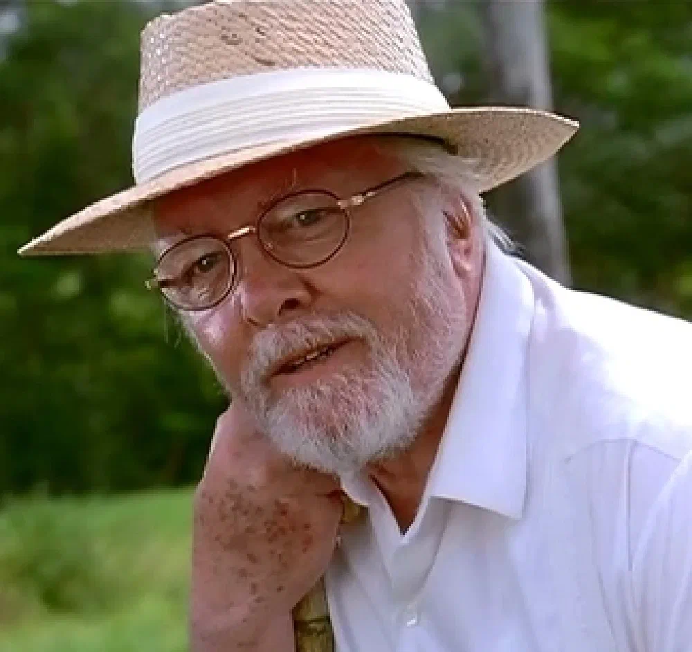 Richard Attenborough as John Hammond from Jurassic Park, an older man with a white beard wearing a straw boater hat and round wire-rim glasses.