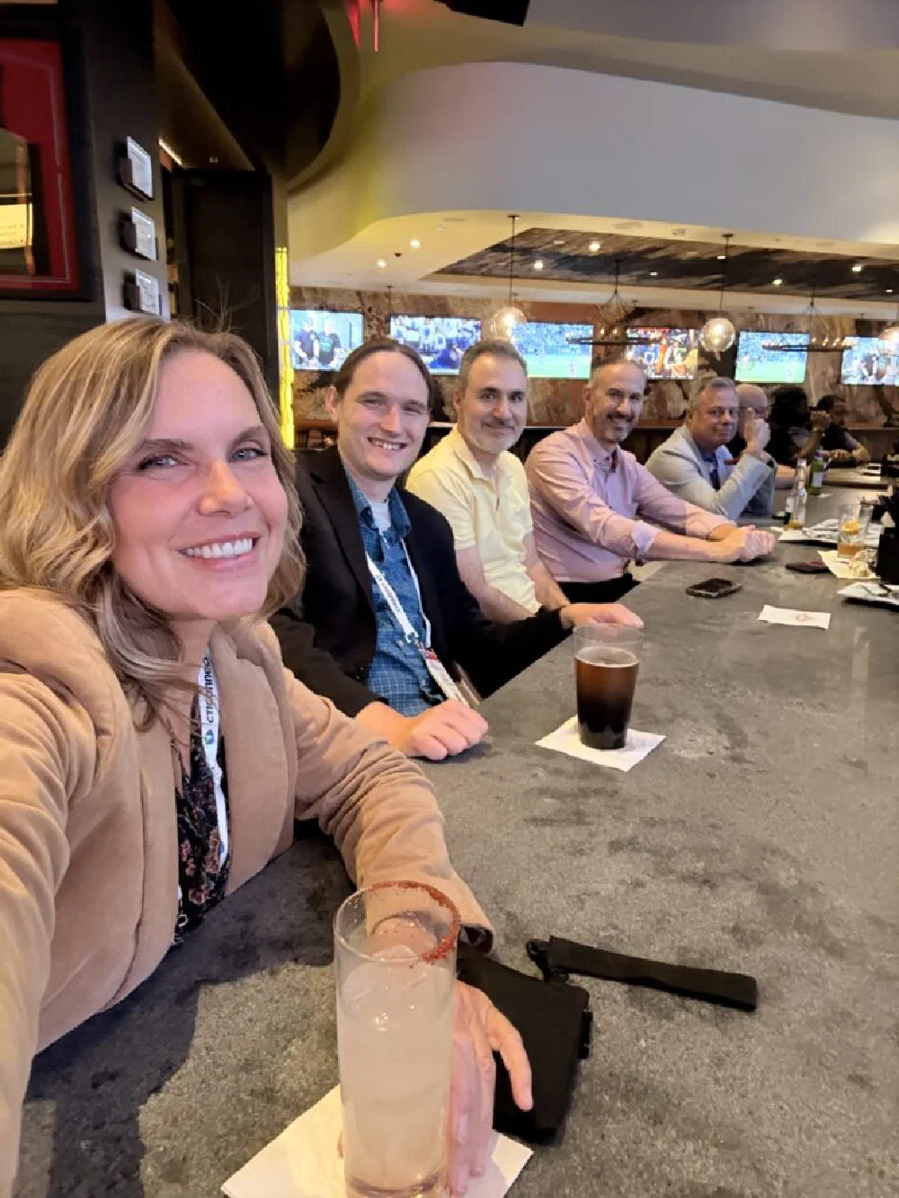 Conference selfie of a smiling woman in a tan blazer at a sports-bar table, with five men in business attire seated alongside her.