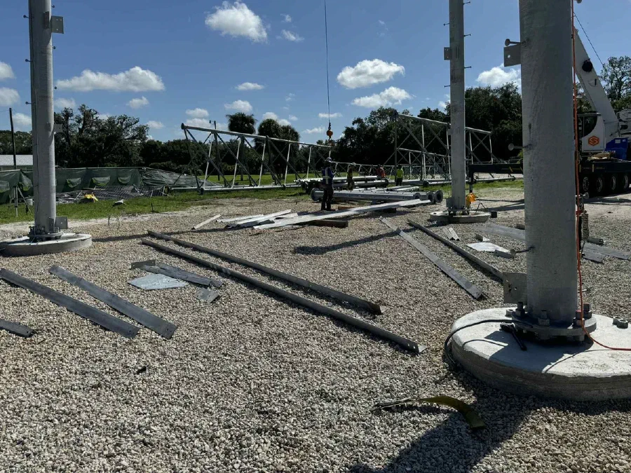 A Vertical Axis crew standing steel on a new monopole build: pole sections on the ground, a crane line overhead, and workers in hard hats among the foundations.