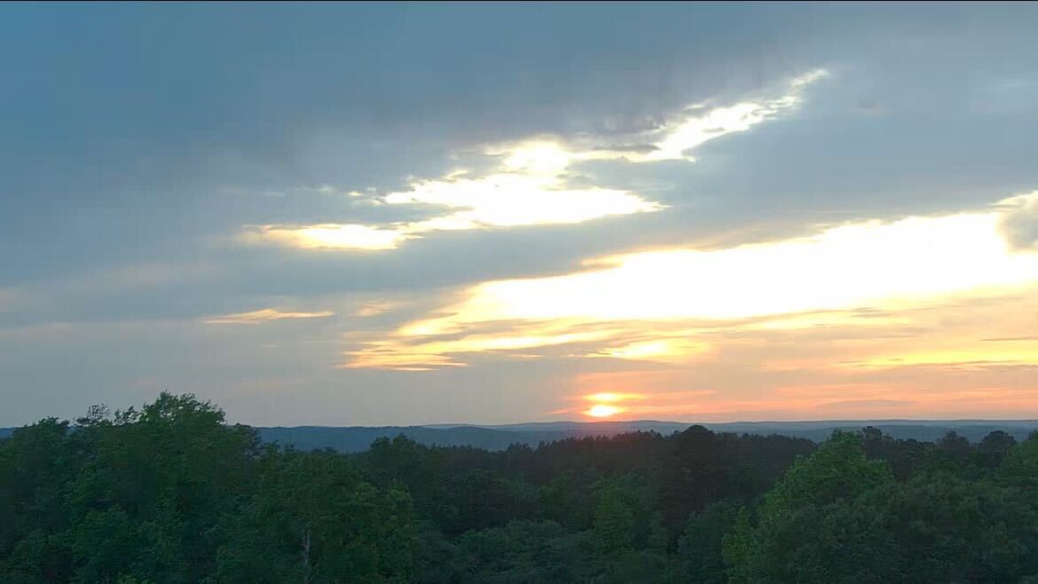 Wide-angle sunset photo of central Alabama, with the sun dropping behind low forested ridges under streaked orange and gray clouds.